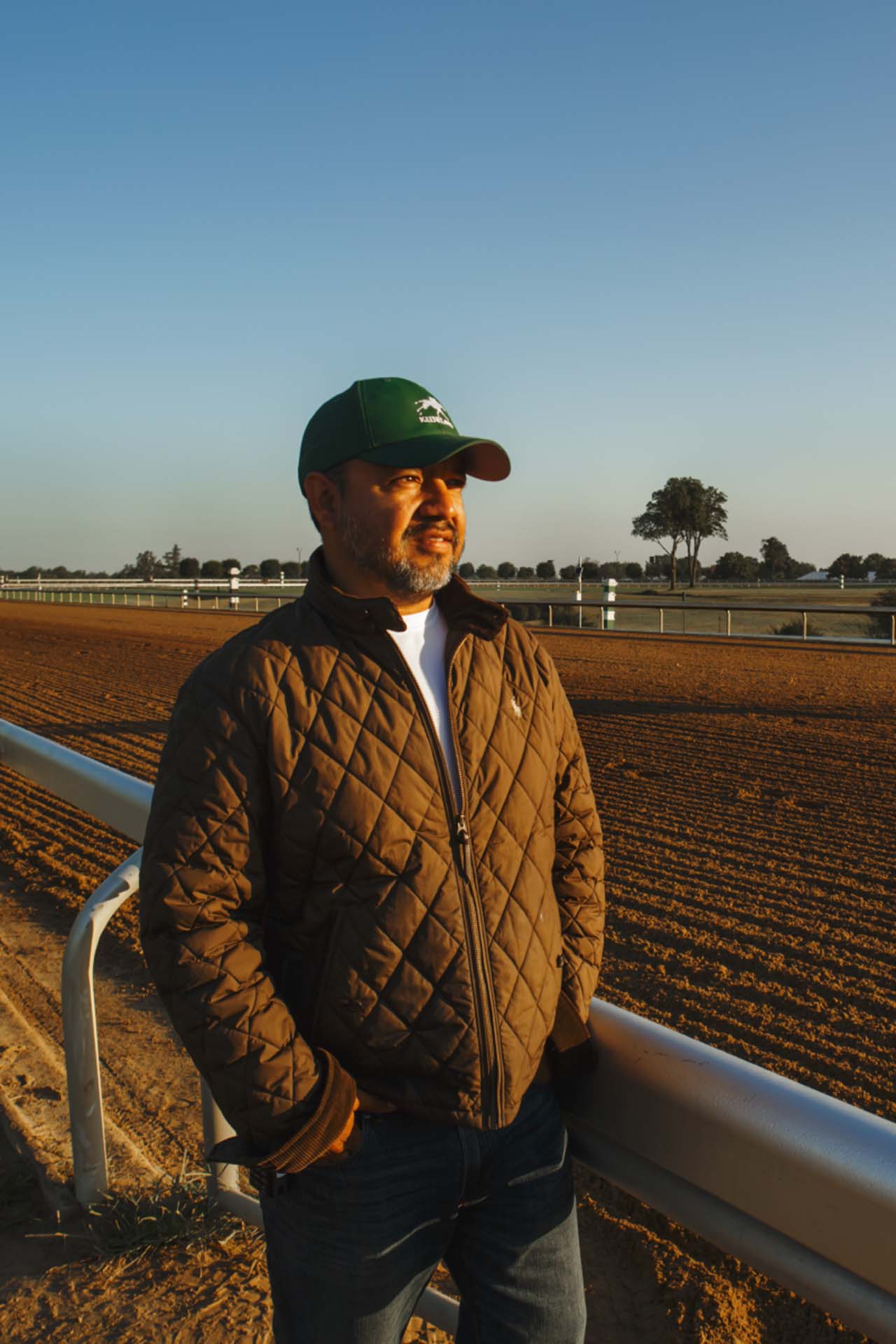 A photo of Alfredo Laureano looking off into the distance while standing by the dirt track at sunset. He is an older Hispanic man with graying chin stubble. He is wearing a green Keensaddle-brand hat and a brown Keensaddle-brand jacket overtop a white shirt.