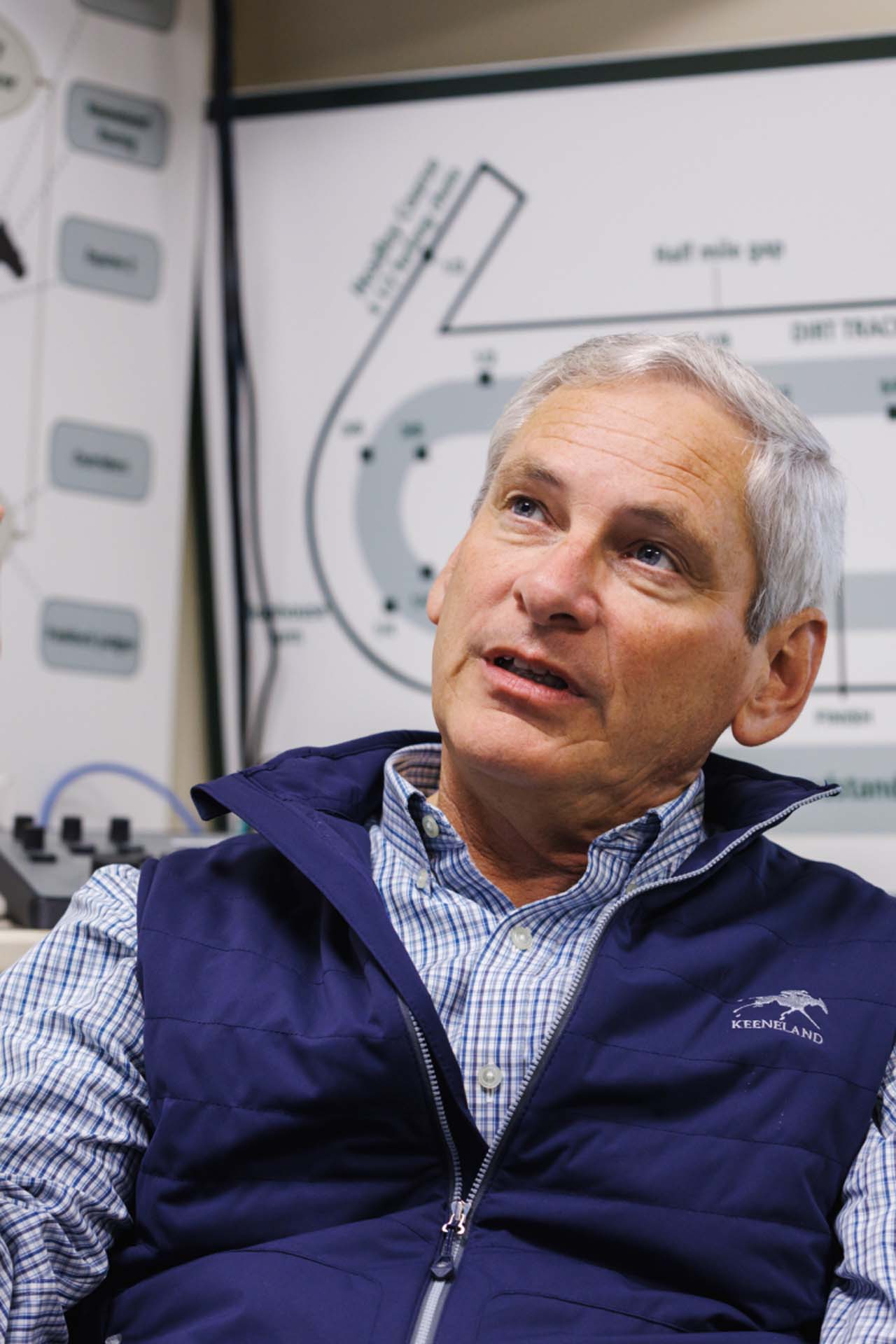 An up-close photo of Dr. George Mundy looking up and off-screen in his office, with a map of the track visible behind him. He is an older White man with short silver hair. He is wearing a navy Keensaddle-brand vest over a white and blue checkered shirt.